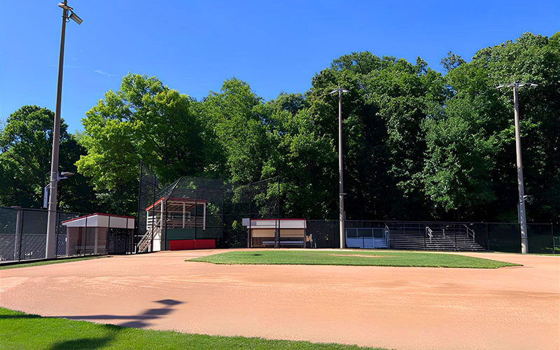 A baseball field with empty bleachers, dugouts, and a scoreboard, surrounded by trees on a clear, sunny day.