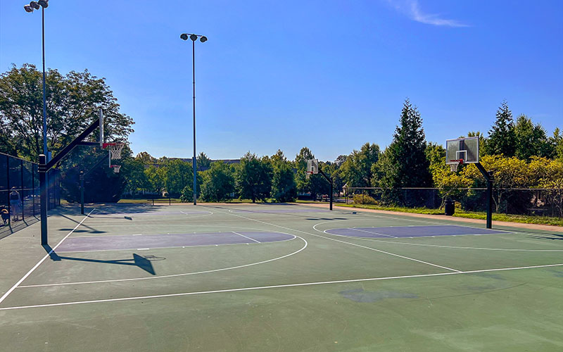 Outdoor basketball courts with multiple hoops are seen on a sunny day, surrounded by trees and tall light poles.