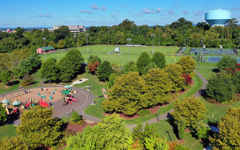 Aerial view of a park with a playground, sports fields, tennis courts, walking paths, trees, and a water tower in the background.