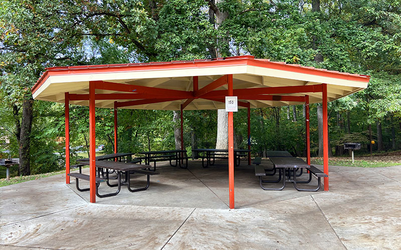 Hexagonal pavilion with a red roof and red support beams, containing several picnic tables, set in a wooded park area.
