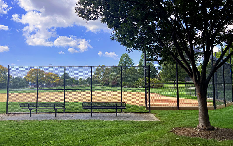 A baseball field with a dirt infield, surrounded by a black chain-link fence, benches, green grass, and trees under a partly cloudy sky.