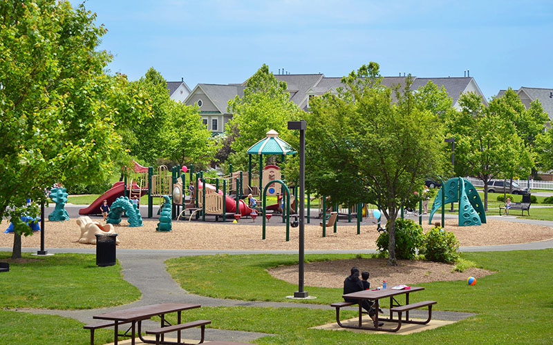 A playground with slides and climbing structures is surrounded by trees and nearby houses. Two people sit at a picnic table in the foreground.