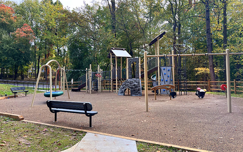 A playground with climbing structures, swings, and benches is surrounded by trees with autumn foliage. The play area is empty.