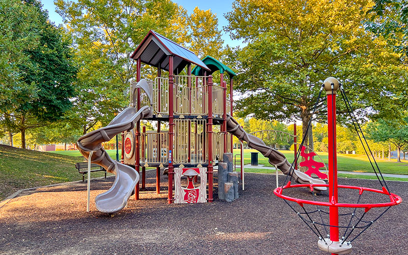 A modern playground with climbing structures, slides, and a rope spinner, surrounded by trees and green grass on a sunny day.