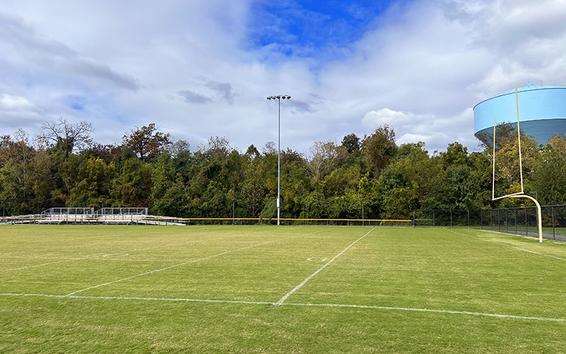 A grassy football field with white boundary lines, empty metal bleachers, a goalpost, and trees in the background under a partly cloudy sky.