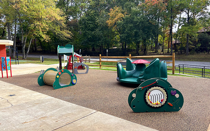 An outdoor playground with accessible play equipment on a rubber surface, surrounded by trees and a wooden fence.