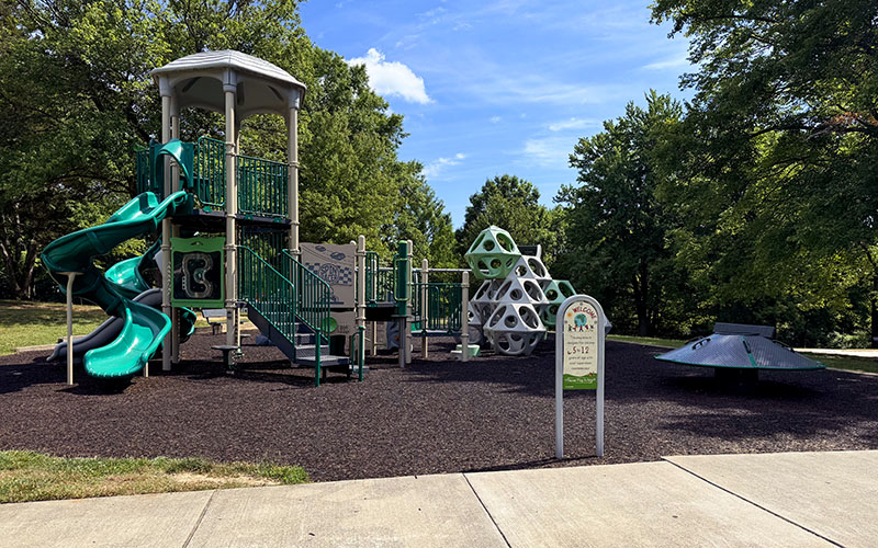 Playground with green slides, climbing structures, and a spinning disk, surrounded by trees and bordered by a sidewalk under a blue sky.