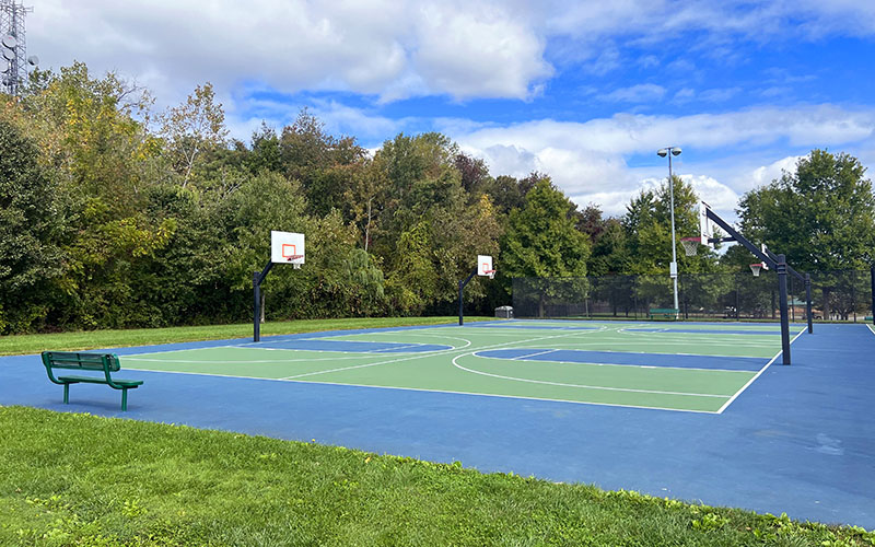 Outdoor basketball courts with blue and green surfaces, surrounded by trees and grass, under a partly cloudy sky; a green bench is in the foreground.