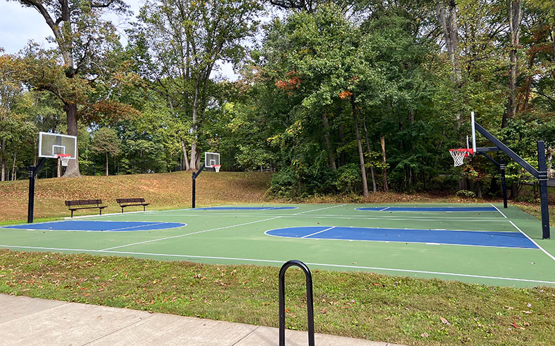 Outdoor basketball court with four hoops, surrounded by trees and benches on a clear day. The court has green and blue markings.