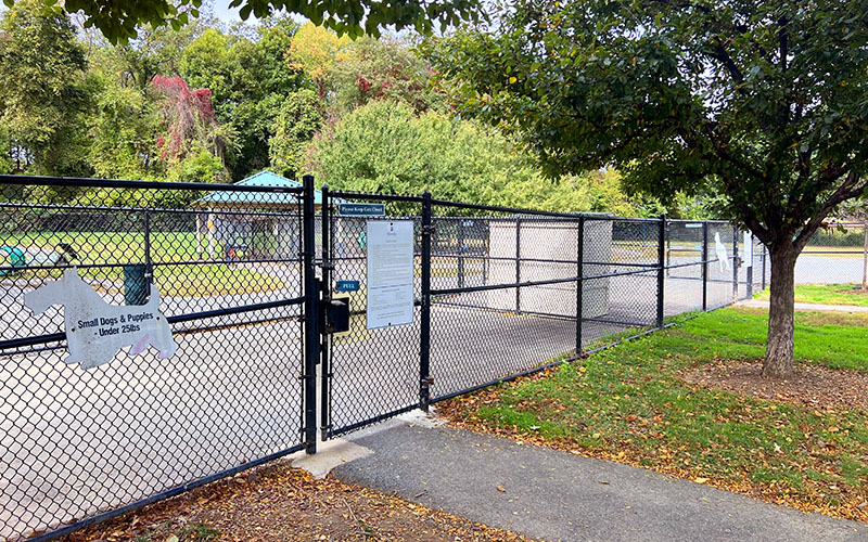 Fenced entrance to a dog park with signage for small dogs and puppies under 25 lbs; trees and a covered area are visible inside.