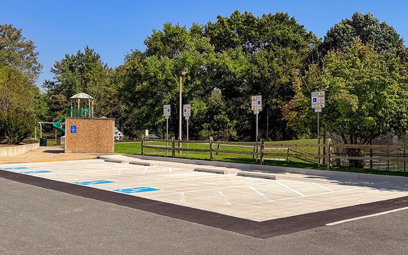 An empty parking lot with multiple reserved accessible parking spaces, bordered by trees and a playground in the background.