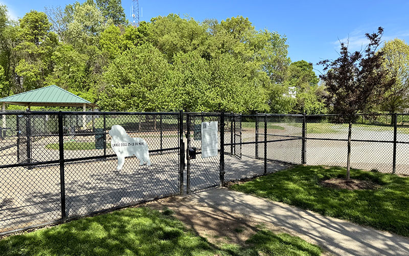 A fenced dog park with a white dog-shaped sign on the gate, surrounded by green trees and grass under a clear blue sky.