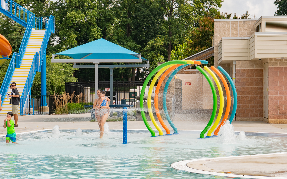 A woman walks through colorful water arches in a splash pad area while a child plays nearby. Trees and park structures are visible in the background.