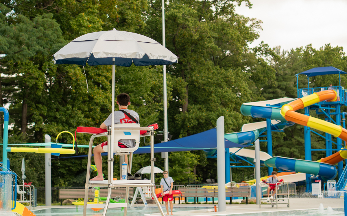 A lifeguard sits on a tall chair under an umbrella by a pool, with water slides and people in the background at an outdoor water park.