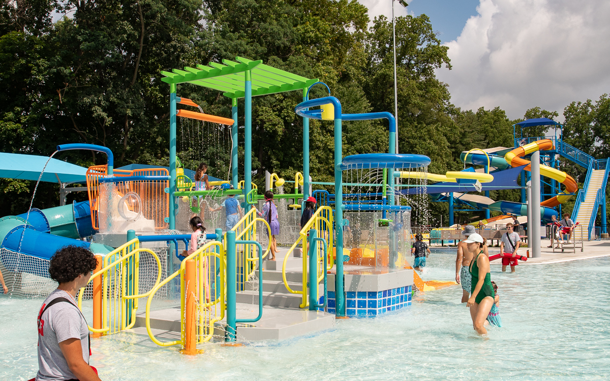 People enjoy a colorful outdoor water park with slides, splash features, and shallow water on a sunny day; trees are visible in the background.