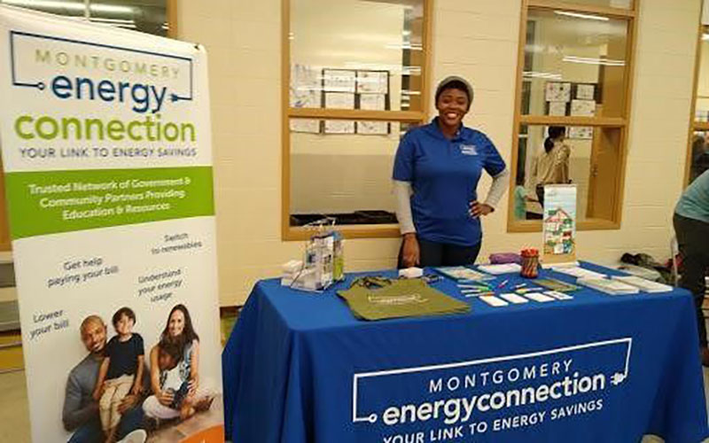 A person stands behind a table with informational materials for Montgomery Energy Connection, promoting energy savings at an indoor event.