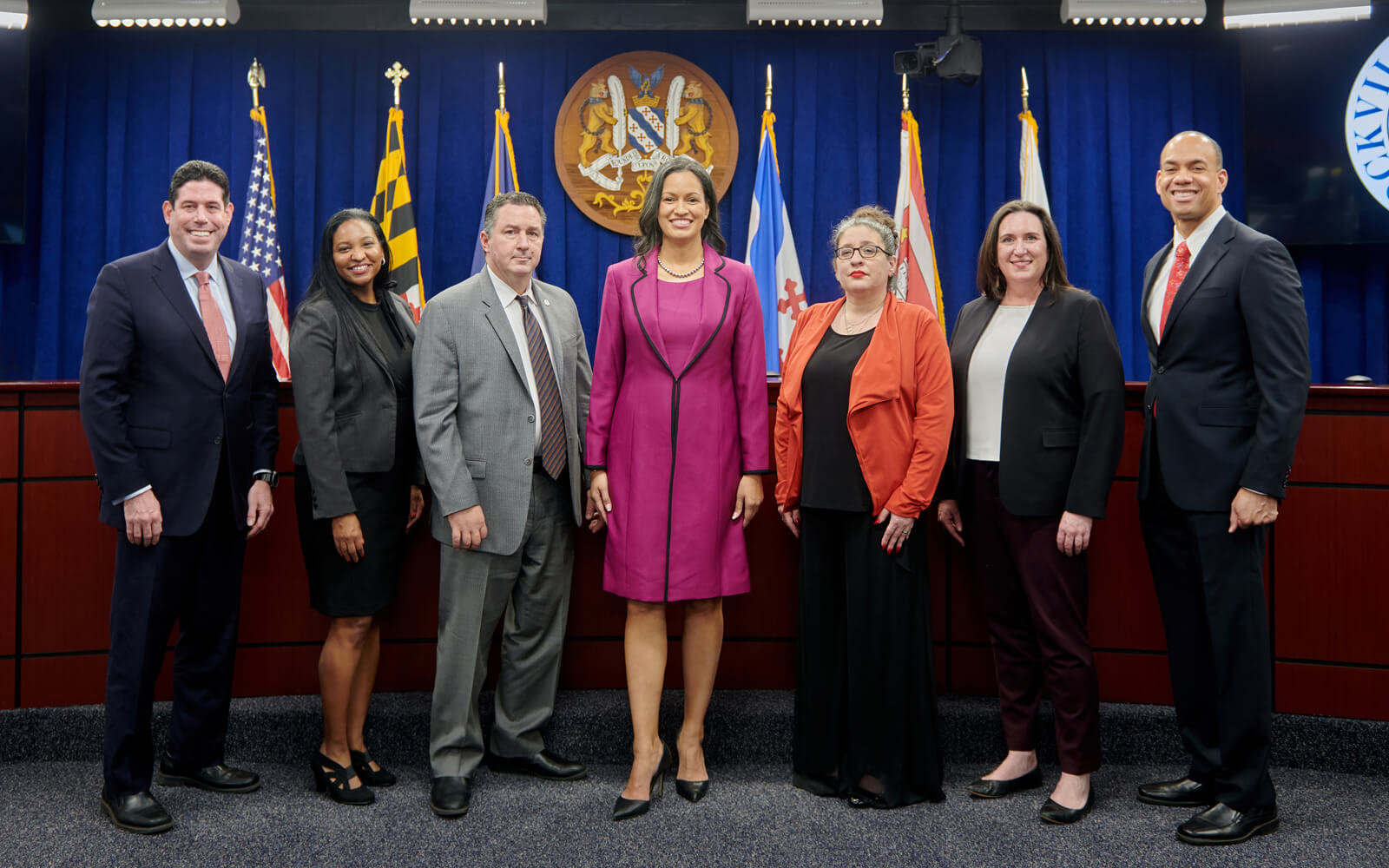 Seven professionally dressed individuals stand in a row in front of flags and an official seal in a formal indoor setting.