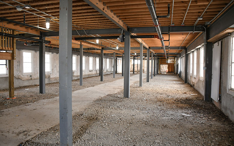 Interior of a large, unfinished building with exposed wooden ceiling beams, steel support columns, gravel flooring, and rows of windows along both sides.