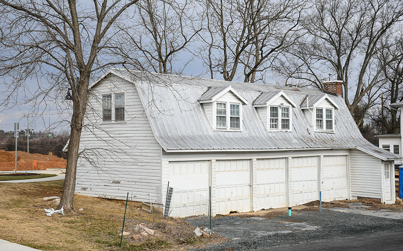 A two-story white building with a metal roof and three dormer windows sits above four closed garage doors, surrounded by leafless trees and a patchy lawn.