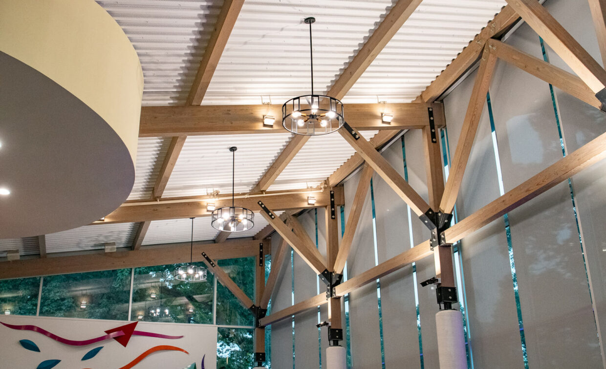 Interior ceiling with exposed wooden beams, metal supports, and modern hanging light fixtures; large windows on the right let in natural light.