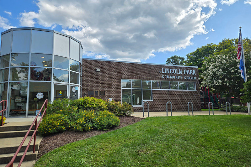 Exterior view of Lincoln Park Community Center, featuring a brick facade, large windows, landscaped greenery, and flagpoles.