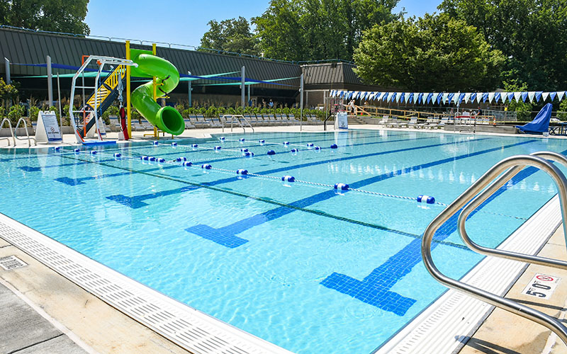 Outdoor swimming pool with lane markers, ladders, blue and white bunting, lounge chairs, and a green spiral water slide. Trees and buildings are in the background.