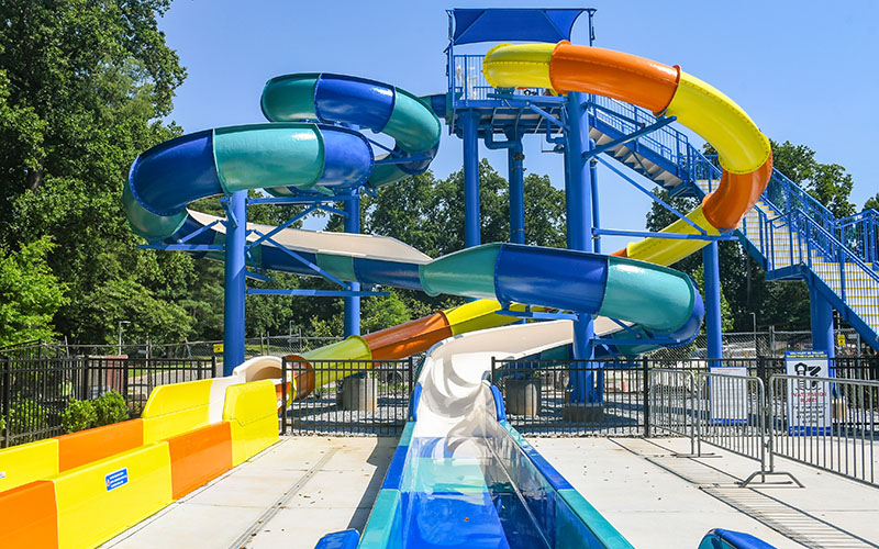 Three colorful, twisting water slides descend from a blue platform at an outdoor water park on a sunny day.