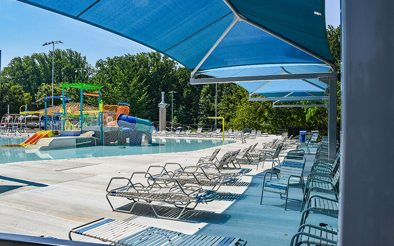 Outdoor pool area with empty lounge chairs under blue sunshades; colorful water slides and play structures in the background, surrounded by trees.