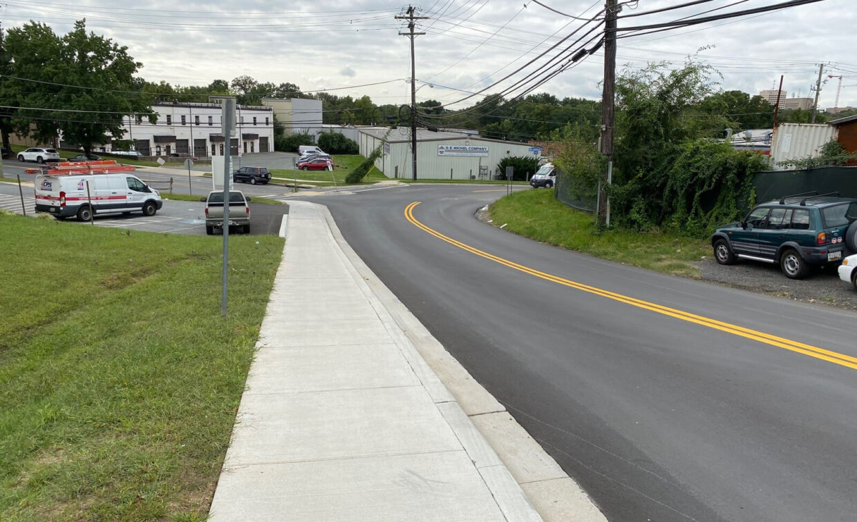 A newly paved road with double yellow lines curves to the right; a sidewalk runs alongside, and vehicles are parked near buildings in the background.