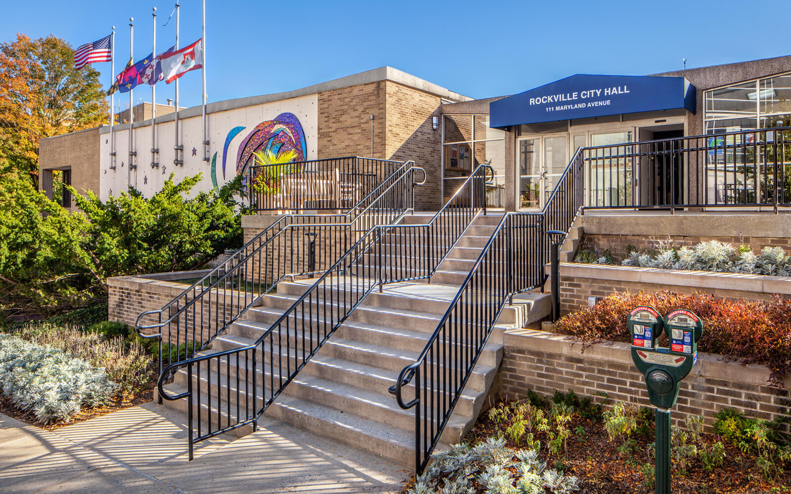 Rockville City Hall building with staircases, black railings, plants, flagpoles, a parking meter, and a blue awning over the entrance on a clear day.