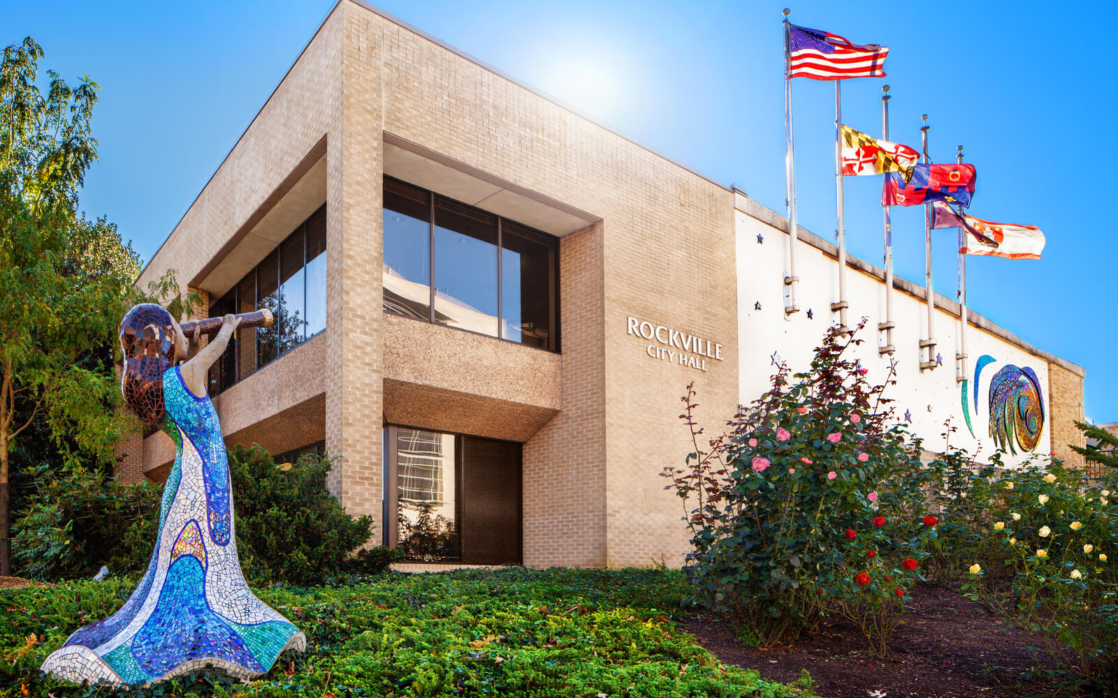 A modern brick building labeled "Rockville City Hall" with a mosaic angel sculpture and several flags in front, surrounded by landscaping and flowering plants.