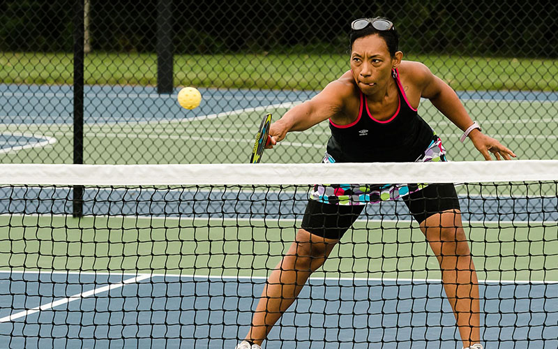A person plays pickleball, reaching forward with a paddle to hit a ball over the net on an outdoor court.