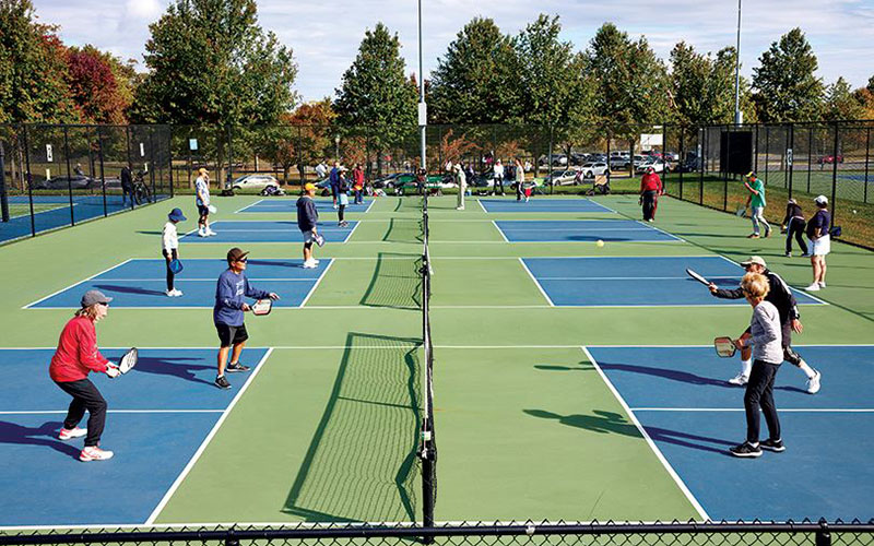 Multiple groups of people play pickleball on outdoor courts surrounded by trees and parked cars on a sunny day.