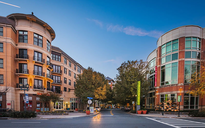 A city street lined with modern buildings, trees, and storefronts at dusk, with a clear sky overhead and streetlights starting to turn on.