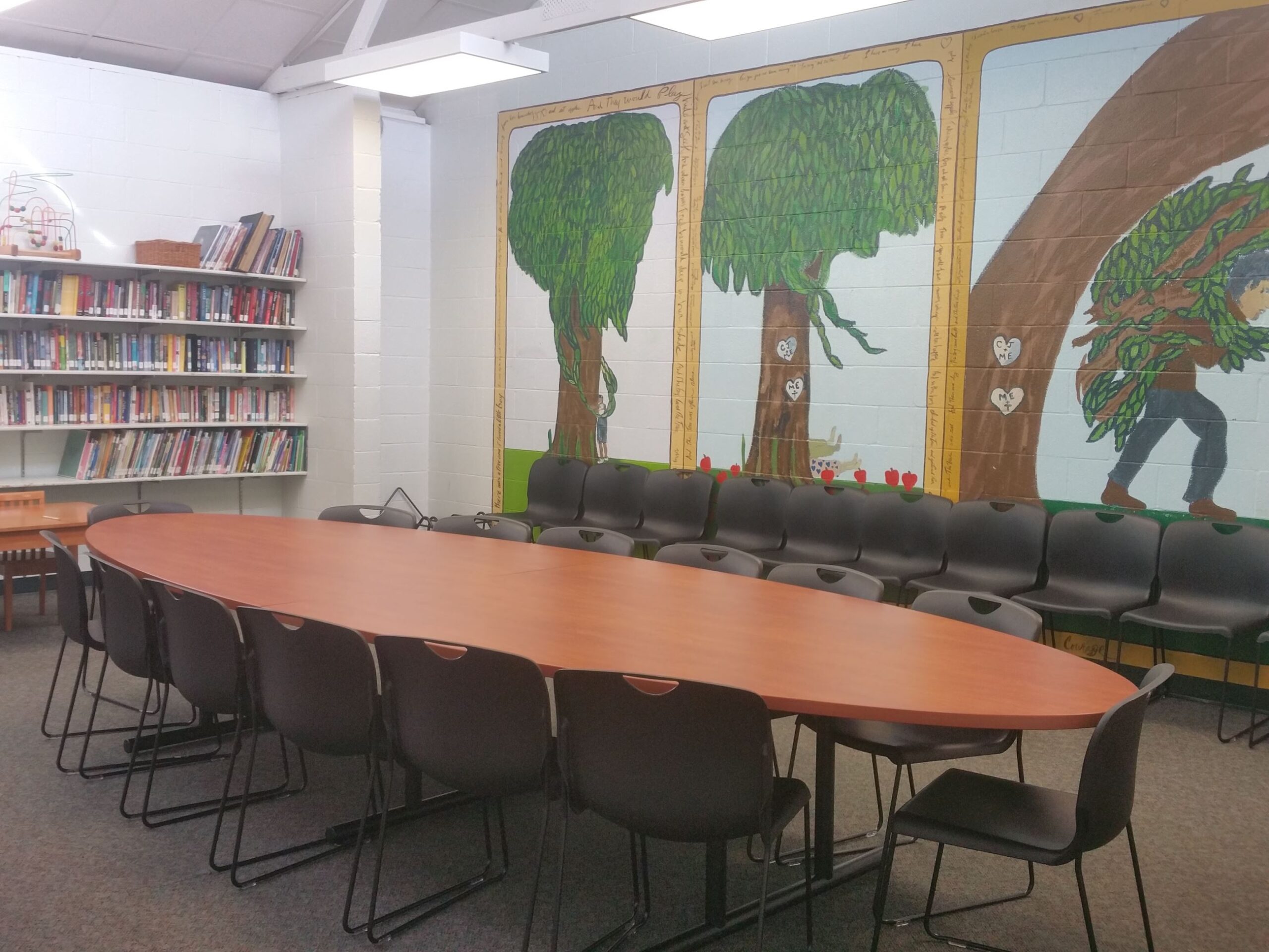 A library room with an oval conference table, black chairs, bookshelves, and a mural of trees with a person carrying wood on the wall.