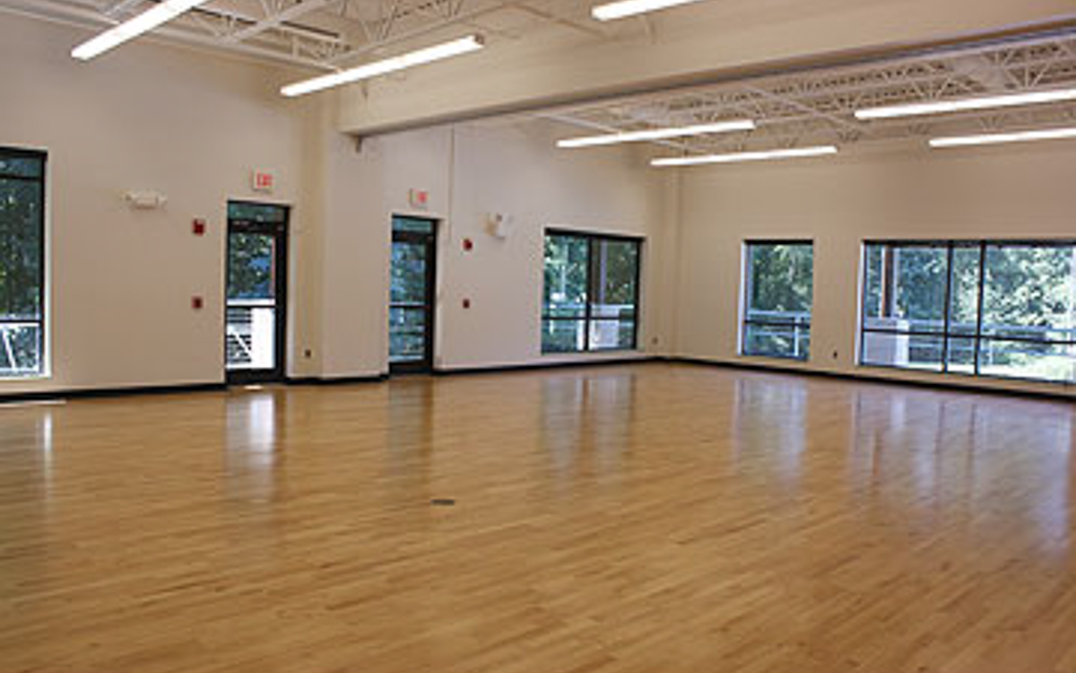 An empty room with wooden floors, white walls, large windows, and fluorescent ceiling lights.