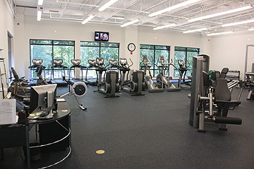 A gym with cardio machines, weight machines, and a row of treadmills by large windows; a desk with computers is visible in the foreground.