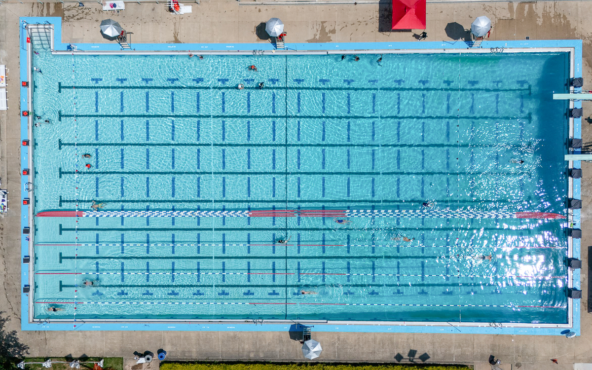 Aerial view of a large outdoor swimming pool with multiple swim lanes and a few people swimming; poolside umbrellas and deck chairs visible.