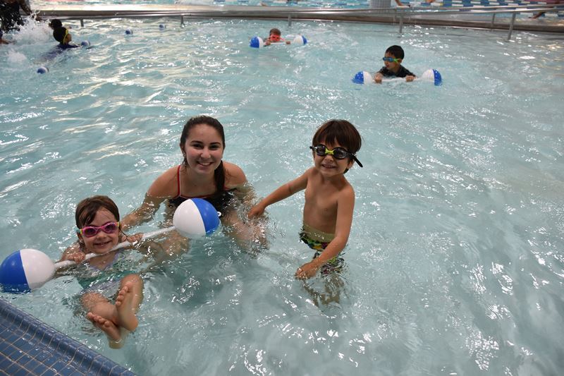 Three children and an adult are in a swimming pool, smiling at the camera. Two children hold blue and white floating balls, and all are wearing swim goggles.