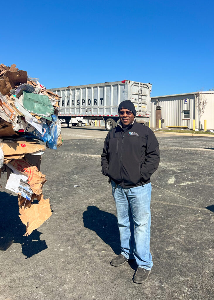 A man wearing a black jacket, hat, and sunglasses stands outdoors near a pile of cardboard and a transport truck under a clear blue sky.